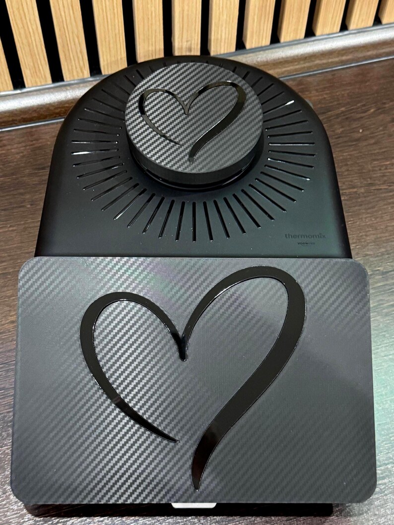 以下が含まれることがあります： カーボンファイバーのハートデザインが施された黒いキッチン家電。ハート型のアクセントが付いた丸い上部と、お揃いのハートが付いた長方形のベースがあります。「Thermomix」のブランド名がベースに表示されています。