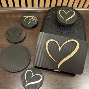 May include: A black carbon fiber design with a heart-shaped cutout and a wooden heart design. The image includes a black appliance with a heart design, along with several coasters with heart and initial designs.