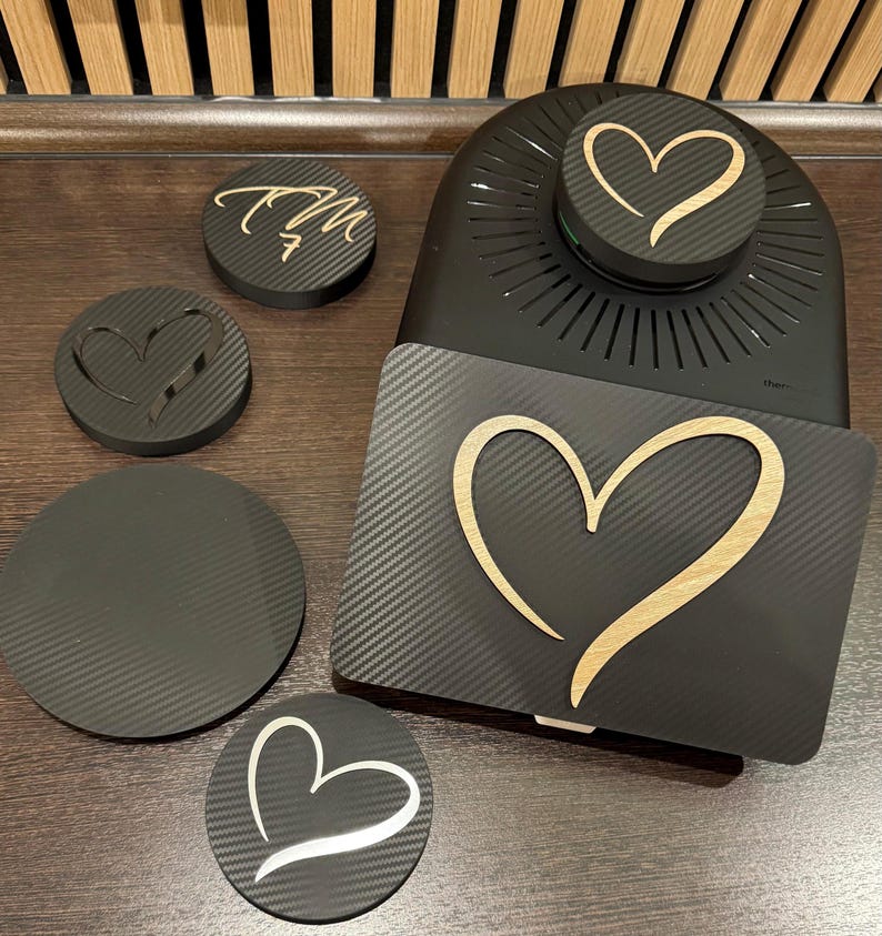 May include: A black carbon fiber design with a heart-shaped cutout and a wooden heart design. The image includes a black appliance with a heart design, along with several coasters with heart and initial designs.
