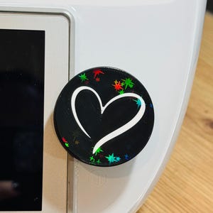Könnte beinhalten: Ein runder schwarzer Handy-Griff mit einem weißen Herz-Design und bunten Glitzer-Akzenten. Der Griff ist an einem weißen Gerät mit dem Wort "thermomix" angebracht.