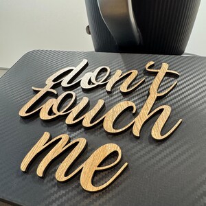 May include: Wooden sign with the words "don't touch me" in a cursive font. The sign is made of light-colored wood and is placed on a black surface. A black and silver object is in the background.