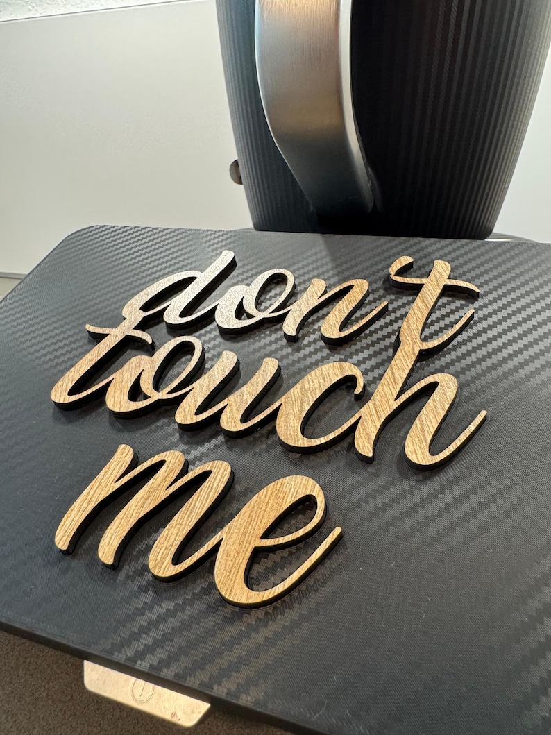 May include: Wooden sign with the words "don't touch me" in a cursive font. The sign is made of light-colored wood and is placed on a black surface. A black and silver object is in the background.