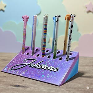 Könnte beinhalten: Ein violetter Glitzer-Stiftehalter mit dem Namen Johanna in schwarzer Schreibschrift auf der Vorderseite. Der Halter hat mehrere kreisförmige Schlitze, in denen verschiedene Stifte mit Tier- und Figurenaufsätzen stecken, darunter ein Schmetterling, ein Panda und eine Kuh.