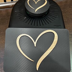 May include: Black kitchen appliance with a carbon fiber texture and a wooden heart design. The appliance has a round top with a heart-shaped design and a rectangular base with a larger heart. The word "thermomix" is printed on the base.