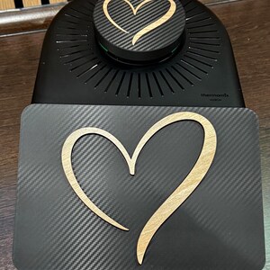 以下が含まれることがあります： カーボンファイバーの質感と木製のハートのデザインが施された黒いキッチン家電。家電にはハートのデザインが施された丸い上部があり、「thermomix」の文字が見えます。木製のハートは明るい茶色です。