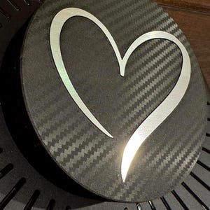 May include: A round, black object with a carbon fiber texture. A silver heart shape is centered on the object. The heart has a reflective, metallic appearance. The object is slightly angled, revealing a glimpse of a dark, textured surface.