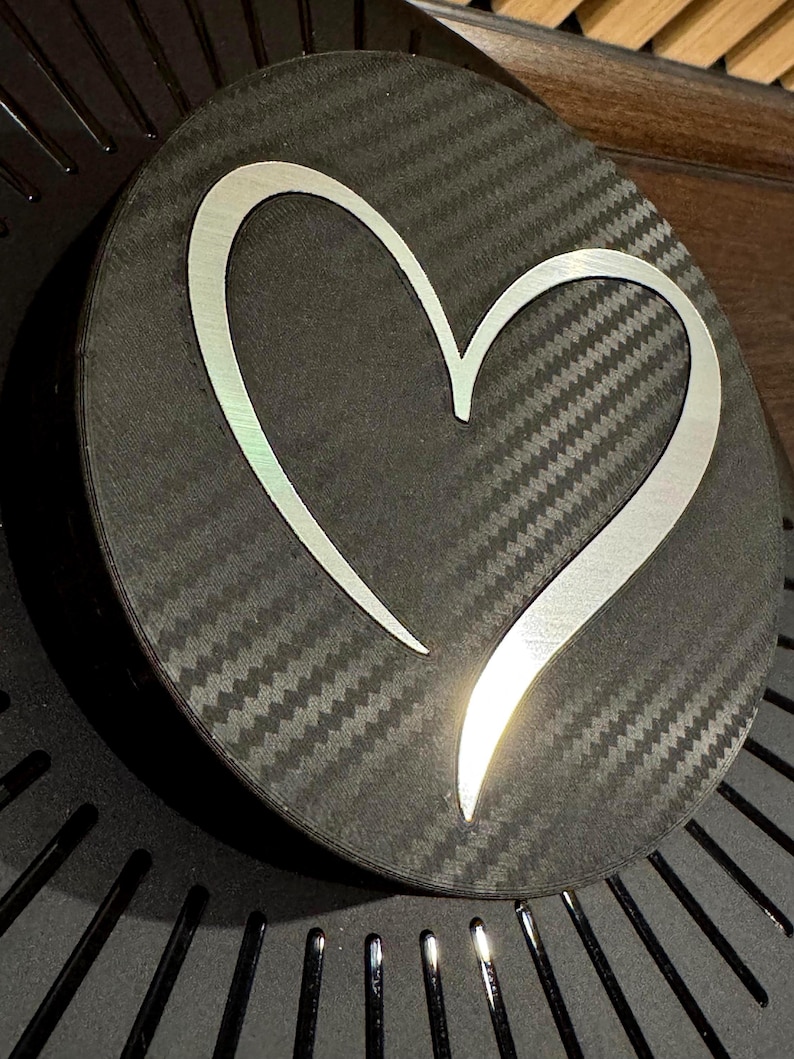 May include: A round, black object with a carbon fiber texture. A silver heart shape is centered on the object. The heart has a reflective, metallic appearance. The object is slightly angled, revealing a glimpse of a dark, textured surface.