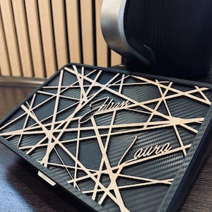 May include: A black and gray rectangular case with a geometric wooden overlay. The overlay features the name "Adrian aura" in a stylized font. The case is set against a wooden background and a black object with a silver trim.