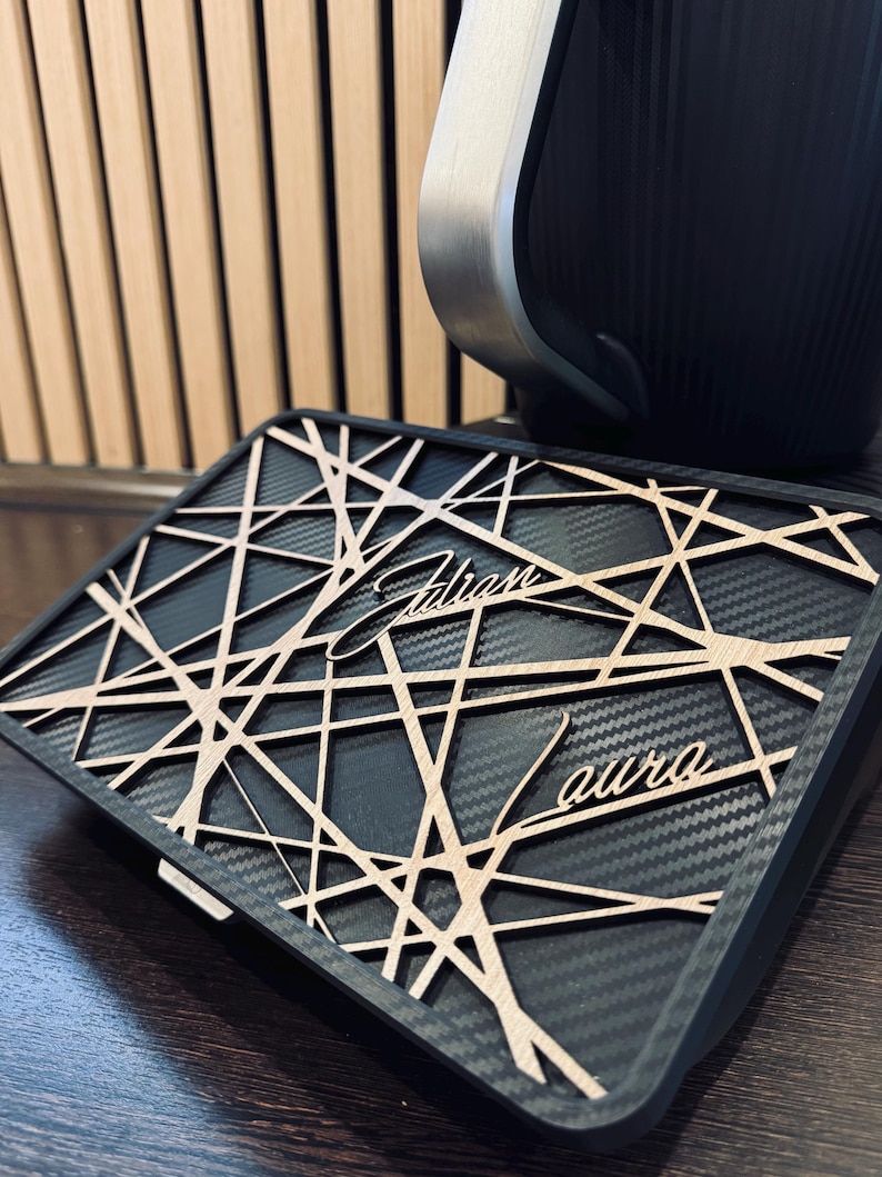 May include: A black and gray rectangular case with a geometric wooden overlay. The overlay features the name "Adrian aura" in a stylized font. The case is set against a wooden background and a black object with a silver trim.