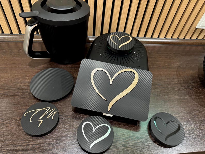 May include: A black coffee mug and several black coasters with heart and initial designs. The coasters have a textured surface and a wooden heart design. The mug has a silver handle.