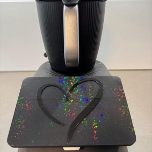 May include: Black electric kettle with a silver handle and a black base. A black tray with a heart-shaped cutout and colorful starburst pattern is in front of the kettle.