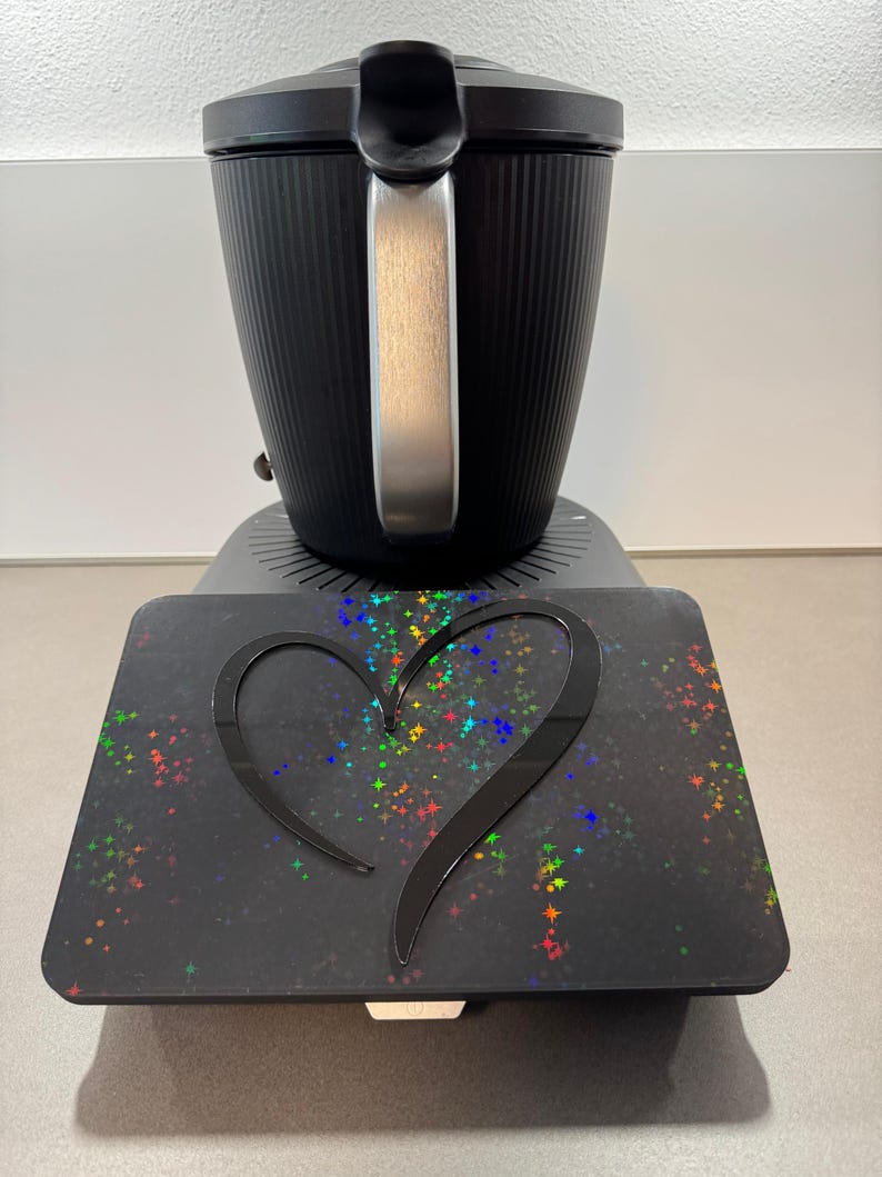 May include: Black electric kettle with a silver handle and a black base. A black tray with a heart-shaped cutout and colorful starburst pattern is in front of the kettle.