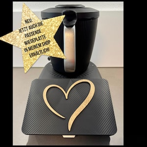 Könnte beinhalten: Schwarzes Küchengerät mit silbernem Griff und schwarzem Sockel. Ein schwarzes rechteckiges Tablett mit einem Holzherz-Design befindet sich vor dem Gerät. Eine goldfarbene sternförmige Grafik mit deutschem Text befindet sich in der oberen linken Ecke.