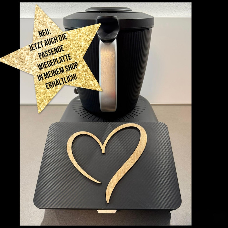May include: Black kitchen appliance with a silver handle and a black base. A black rectangular tray with a wooden heart design is in front of the appliance. A gold star-shaped graphic with German text is in the upper left corner.