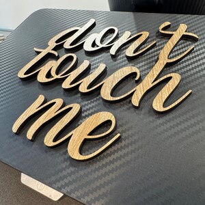 以下が含まれることがあります： 筆記体の「don't touch me」という文字が書かれた木製のサイン。文字は明るい茶色の木でできており、濃い灰色の表面に置かれています。