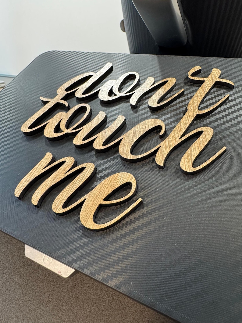 以下が含まれることがあります： 筆記体の「don't touch me」という文字が書かれた木製のサイン。文字は明るい茶色の木でできており、濃い灰色の表面に置かれています。