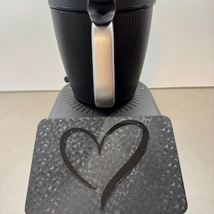 May include: A black coffee maker with a silver handle and a textured, ribbed design. It sits on a gray base with a decorative heart-shaped accent. The base has a geometric pattern.