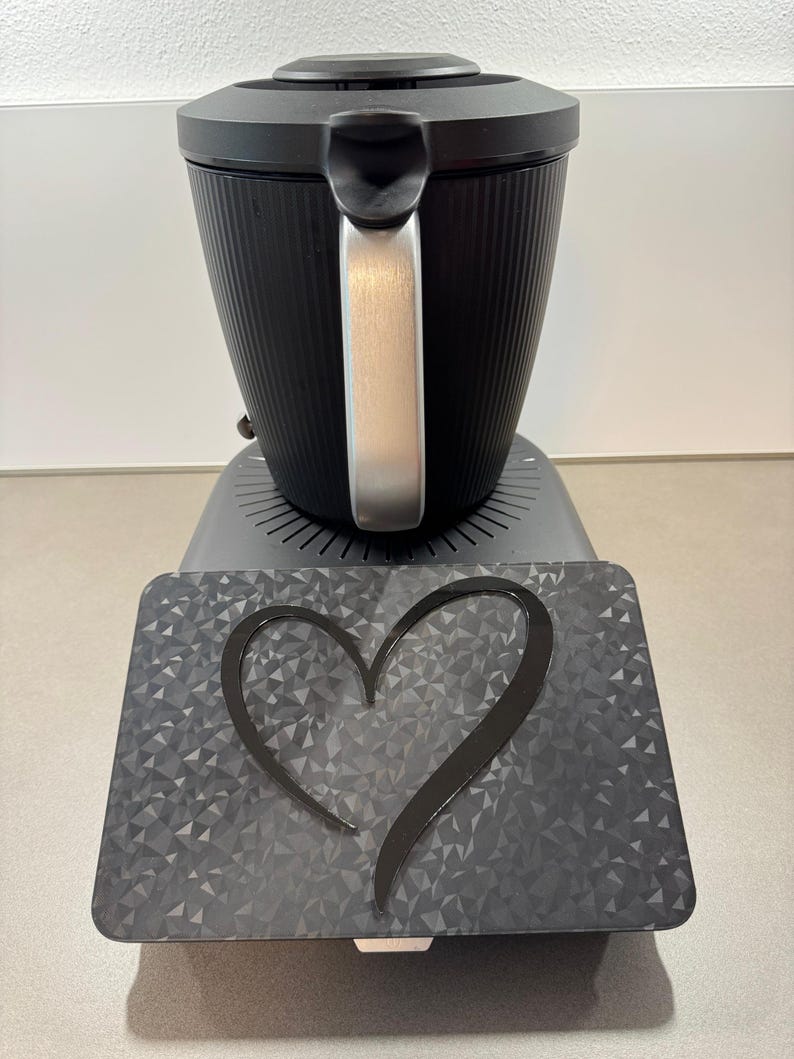 May include: A black coffee maker with a silver handle and a textured, ribbed design. It sits on a gray base with a decorative heart-shaped accent. The base has a geometric pattern.
