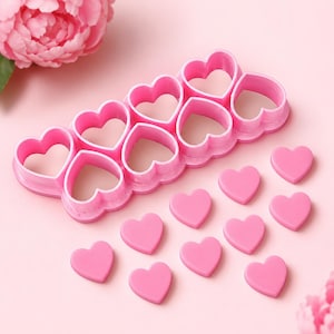May include: A pink plastic multi-heart cookie cutter tool featuring a connected frame of eight heart-shaped molds, accompanied by ten separate small pink heart-shaped cutouts arranged in a scattered pattern on a light pink surface.