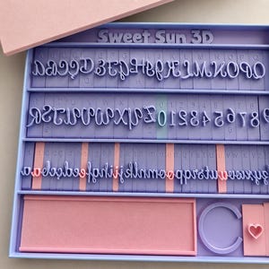 May include: A light purple and pink Sweet Sun 3D stamp set with letters, numbers, and symbols. The set includes a tray with a pink rectangular insert, a purple C-shaped stamp, and a pink heart-shaped stamp.