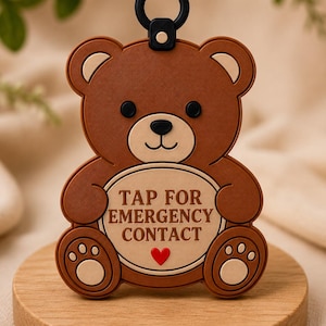 May include: A brown bear-shaped silicone tag with a black circular loop at the top. The bear has a cream-colored belly patch featuring the text TAP FOR EMERGENCY CONTACT in brown letters and a small red heart icon below the text.