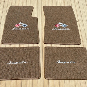 May include: A set of four brown carpeted car floor mats. The two front mats feature an embroidered Impala logo with a red and white chequered flag design. The two rear mats feature the word Impala embroidered in white script lettering.