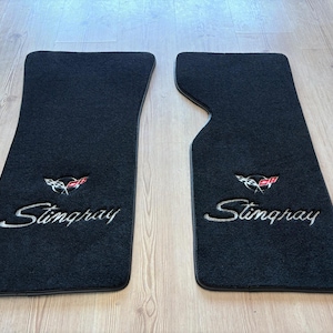 May include: A pair of black automotive floor mats featuring the embroidered Stingray script and the crossed-flags emblem in red, white, and black. The mats have a textured carpet surface and are shaped to fit specific vehicle footwells.