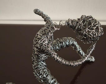 May include: A handcrafted metal wire sculpture depicting a kneeling figure holding a spherical object. The sculpture is made of silver-colored wire, with intricate details and a reflective surface. The artwork is a unique piece of home decor.