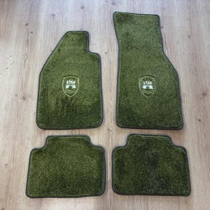 May include: A set of four olive green automotive floor mats with a textured, carpeted surface and dark trim. Two of the mats feature a small, embossed shield-shaped emblem in the center, while the other two mats are plain rectangular shapes.