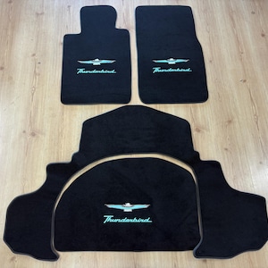 Puede incluir: Un juego de cuatro alfombrillas negras para coche con la palabra Thunderbird en letra cursiva turquesa y un emblema de pájaro estilizado en turquesa y blanco bordado en cada pieza. El juego incluye dos alfombrillas delanteras y dos traseras.