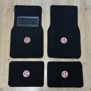 Puede incluir: Un juego de cuatro alfombrillas negras para coche con un parche circular bordado que muestra un logotipo rojo y blanco con el texto Racing Team Dodge. La alfombrilla del conductor incluye un refuerzo negro con textura.
