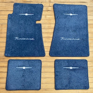 Puede incluir: Un juego de cuatro alfombrillas de moqueta azul oscuro para vehículo. Cada alfombrilla presenta un logotipo Thunderbird bordado en blanco y la palabra Thunderbird en cursiva. Las dos alfombrillas delanteras son contorneadas y las traseras son cuadradas.