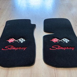 May include: A pair of black automotive floor mats featuring an embroidered emblem of two crossed flags, one red and one checkered, above the red stitched text Stingray. The mats have a contoured shape with a cut-out section on one side.