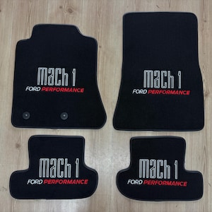 Puede incluir: Un juego de cuatro alfombrillas negras para automóvil con el texto blanco "mach 1" y el texto rojo y blanco "FORD PERFORMANCE" bordados en cada pieza. Las dos alfombrillas delanteras incluyen ojales circulares de montaje.