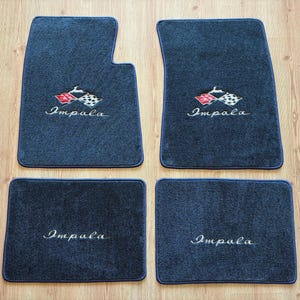 May include: A set of four dark grey automotive floor mats with blue trim. The two front mats feature an embroidered Impala logo with a red and white checkered flag emblem. The two rear mats feature the word Impala embroidered in white script.