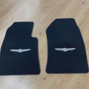 Puede incluir: Dos alfombrillas de coche negras con un emblema alado bordado en color plata centrado en cada pieza. Las alfombrillas tienen una forma contorneada diseñada para el interior de un vehículo, con una textura de moqueta suave y bordes rematados.