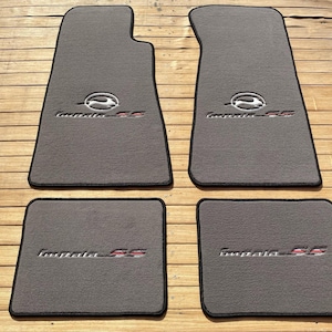 Könnte beinhalten: Ein Set aus vier grauen Automatten mit schwarzer Umrandung. Die zwei größeren vorderen Matten zeigen ein gesticktes Impala SS-Logo mit einem springenden Hirsch-Emblem, während die zwei kleineren hinteren Matten das Impala SS-Textlogo tragen.
