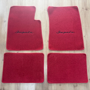 May include: A set of four red automotive floor mats with matching red trim. The two front mats feature a distinct cutout shape and the word Impala embroidered in black script. The two rear mats are rectangular in shape with rounded corners.