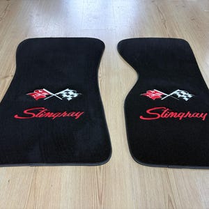 Puede incluir: Dos alfombrillas de coche negras con un logotipo bordado de banderas cruzadas a cuadros y rojas sobre el texto rojo Stingray. Las alfombrillas tienen una forma contorneada con bordes negros y una textura de superficie de alfombra suave.