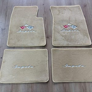 Könnte beinhalten: Ein Set aus vier beigen Teppich-Fußmatten für ein Fahrzeug. Zwei Matten zeigen ein gesticktes Emblem mit einer rot-weißen Zielflagge und einer schwarz-weißen Zielflagge, wobei das Wort Impala darunter gestickt ist. Zwei kleinere rechteckige Matten.