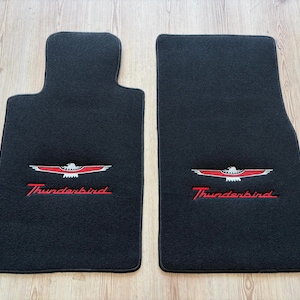 Puede incluir: Dos alfombrillas de coche negras con un emblema Thunderbird bordado en rojo y blanco y la palabra Thunderbird en letra roja. Las alfombrillas tienen una superficie de tela texturizada y están diseñadas para adaptarse al suelo del vehículo.