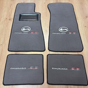 Könnte beinhalten: Ein Set aus vier grauen Automatten mit schwarzer Umrandung. Die beiden vorderen Matten haben eine schwarze Fersenauflage und ein gesticktes Impala SS-Logo mit einem springenden Hirsch. Die beiden hinteren Matten zeigen das gestickte Impala SS-Logo.