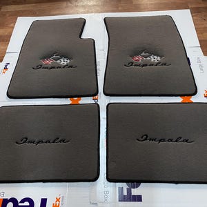 May include: A set of four grey automotive floor mats with black trim. The two front mats feature an embroidered Impala logo with a checkered flag emblem, while the two rear mats feature the embroidered Impala script in a matching grey thread.