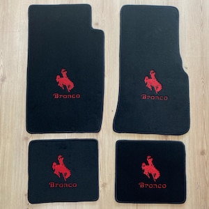 Pode incluir: Um conjunto de quatro tapetes pretos para automóveis com um logótipo bordado a vermelho de um cavalo a empinar e a palavra Bronco em texto vermelho. O conjunto inclui dois tapetes dianteiros maiores com um canto recortado e dois traseiros.