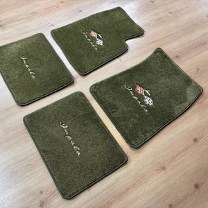 Op de afbeelding: Een set van vier olijfgroene tapijtmatten voor een voertuig. Twee matten zijn voorzien van het geborduurde woord Impala in wit schrift, terwijl de andere twee het woord Impala tonen naast een klein, rood-wit geblokt vlag-embleem.