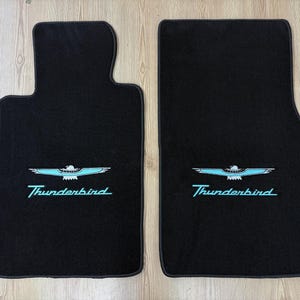 Puede incluir: Dos alfombrillas de coche negras con un logotipo de Thunderbird y texto en cursiva bordados en azul claro. Las alfombrillas tienen una textura de moqueta suave con una forma específica diseñada para el interior de un vehículo.
