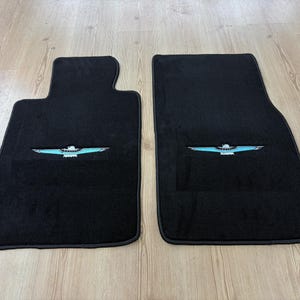 Può includere: Due tappetini per auto neri con un emblema ricamato al centro raffigurante un uccello stilizzato con ali azzurre e corpo bianco. I tappetini hanno una forma rettangolare con un bordo sagomato su un lato, realizzati in materiale di moquette denso.
