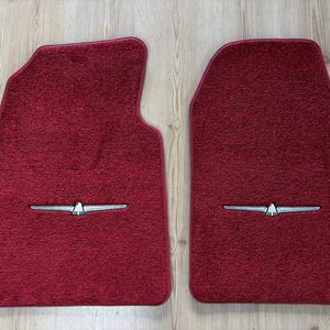 Puede incluir: Un par de alfombrillas de coche de color rojo intenso con una superficie texturizada de moqueta y un emblema bordado en tono plateado de un pájaro estilizado con las alas extendidas en la parte inferior de cada alfombrilla.