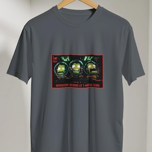 Peut inclure: Un t-shirt gris foncé à manches courtes avec une impression graphique rectangulaire représentant trois personnages verts de l'espace portant des casques. Le motif inclut le texte ADVENTURE BEGINS AT T-MINUS ZERO en lettres rouges.