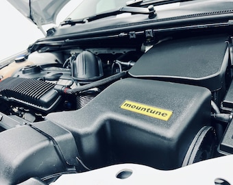 Op de afbeelding: Close-up van een motorruimte met een zwart luchtinlaatsysteem en een gele "mountune" sticker.